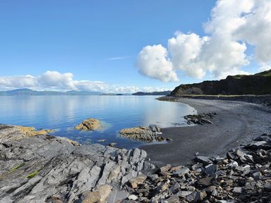 A rocky shoreline with calm water and distant hills under a blue sky at Ardlarach Lodge in Cullipool Isle Of Luing