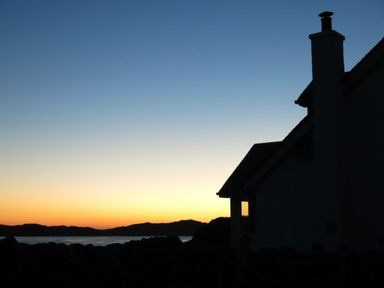 A silhouette of a house with a chimney against a sunset over water and hills at Ardlarach Lodge Cullipool Isle Of Luing