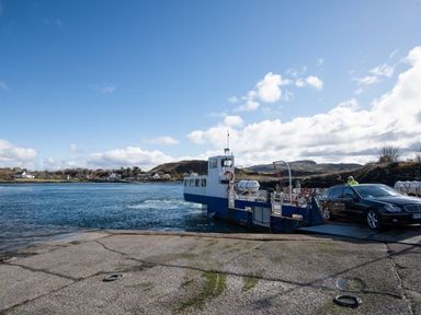 A car boarding a blue and white ferry on a body of water with hills and houses in the background at Ardlarach Lodge in Cullipool Isle Of Luing