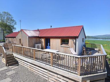 A wooden house with a red roof and blue door with a fenced deck overlooking fields and water at Skylarks Ferintosh near to Dingwall