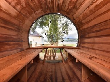 An interior view of a wooden barrel-shaped room with benches on both sides and a round window looking out to a tree and a house at Skylarks in Ferintosh near to Dingwall