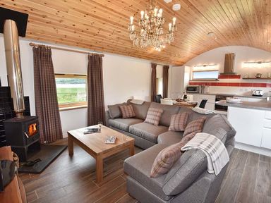 An open living room and kitchen with a gray sectional sofa a wooden coffee table a wood stove and a dining table at Skylarks in Ferintosh near to Dingwall