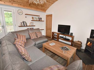 A living room with a gray sectional sofa with plaid cushions a wooden coffee table a flat-screen tv on a wooden stand a wood-burning stove and wall-mounted shelves with books at Skylarks in Ferintosh near to Dingwall