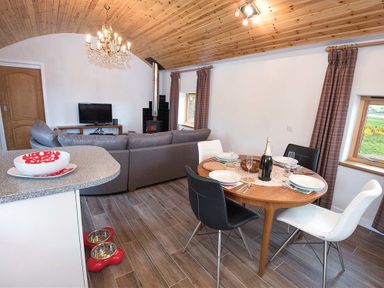 An open-plan living and dining area with a round wooden table set for four black and white chairs a grey sectional sofa a television and a wood-burning stove at Skylarks in Ferintosh near to Dingwall