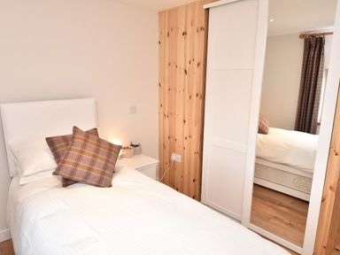 A bedroom with a single bed with white bedding and two plaid pillows next to a white nightstand with a lamp and a sliding wardrobe with a mirror at Skylarks in Ferintosh near to Dingwall