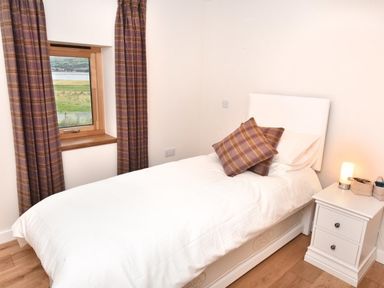 A bedroom with single bed white bedding and two plaid cushions next to window with plaid curtains and a white bedside table with lamp at Skylarks in Ferintosh near to Dingwall