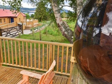A wooden deck with a wooden chair and railing overlooking a grassy area with trees and wooden buildings at Skylarks in Ferintosh near to Dingwall
