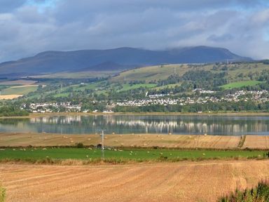 A landscape with fields and hills with a body of water and houses in the distance at Skylarks Ferintosh near to Dingwall