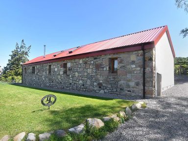 A stone building with a red metal roof and small windows next to a gravel path and a grassy area at Skylarks in Ferintosh near to Dingwall