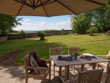 A patio with wooden table and chairs under an umbrella overlooking a garden with a stone wall and trees at Beltie Lodge in Borgue