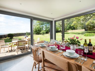 A dining room with a wooden table set for a meal and large windows showing a garden with outdoor seating at Beltie Lodge in Borgue