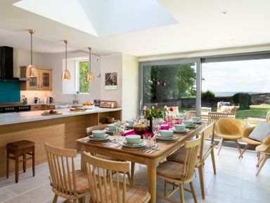 A dining area with a wooden table set for a meal and a kitchen with pendant lights and large glass doors opening to a garden at Beltie Lodge in Borgue