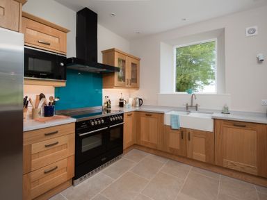 A kitchen with wooden cabinets a black stove a white sink and a window showing trees at Beltie Lodge in Borgue
