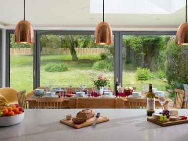 A kitchen island with bread and wine in front of a dining table set with dishes and glasses by large windows showing a garden at Beltie Lodge in Borgue