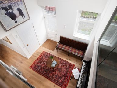 An entryway with a wooden bench with a plaid cushion a piano with sheet music a round wooden table with books on a red patterned rug and a painting of cows on the wall at Beltie Lodge in Borgue