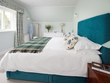A bedroom with a blue bed frame white bedding a green plaid throw two patterned pillows a wooden nightstand with a book a white dresser with flowers and a lamp and patterned curtains at Beltie Lodge in Borgue