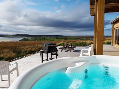 An outdoor wooden deck with a hot tub white chairs a barbecue grill and a picnic table overlooking a grassy landscape and water at Eagle Lodge in Uig near Stornoway