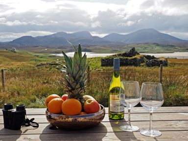 A wooden table outdoors with a bowl of fruit a bottle of wine two empty wine glasses and binoculars with mountains and ruins in the background at Eagle Lodge in Uig near Stornoway
