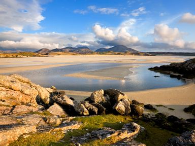 Rocky foreground with patches of grass leading to a sandy beach and a body of water with mountains in the background at Eagle Lodge Uig near Stornoway