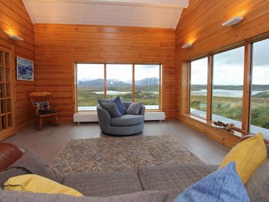 A living room with wood panel walls large windows a rocking chair and upholstered seating at Eagle Lodge in Uig near Stornoway