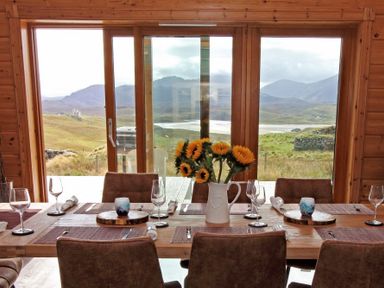 A dining room with a wooden table set with glasses plates and sunflowers in a vase with a view of hills through large windows at Eagle Lodge in Uig near Stornoway