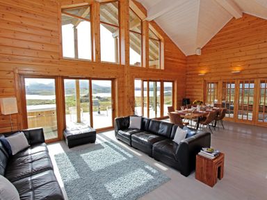 A living room with black leather sofas a blue rug wooden dining table with chairs and large windows at Eagle Lodge in Uig near Stornoway