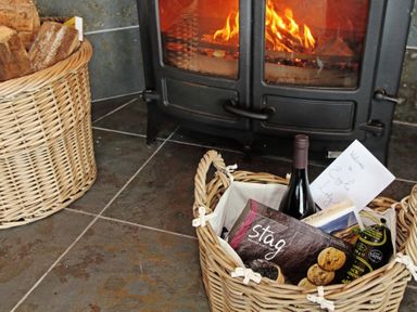 A burning wood stove next to a basket of firewood and a basket with a bottle of wine and snacks at Eagle Lodge in Uig near Stornoway