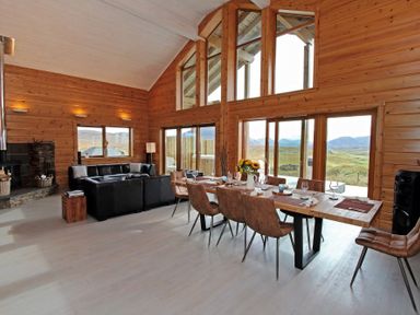 A dining table with chairs and a black sofa in a wooden room with large windows at Eagle Lodge in Uig near Stornoway