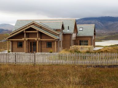 A wooden house with a grey tiled roof behind a wooden fence in a rural area with mountains in the background at Eagle Lodge in Uig near Stornoway