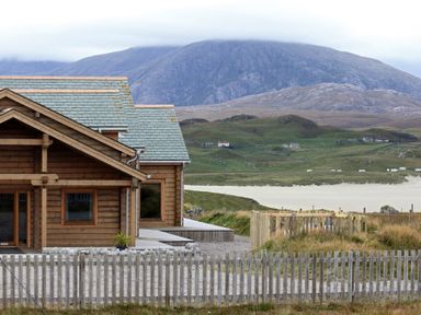 A wooden house with a fenced yard near a beach and hills at Eagle Lodge in Uig near Stornoway