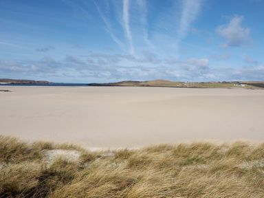 A sandy beach with dunes in the foreground and hills with scattered houses in the background at Eagle Lodge in Uig near Stornoway