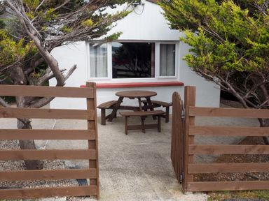 An outdoor area with a round wooden picnic table and benches behind a wooden gate and fence in front of a white building with a window Braemore Chalet Tarbert Isle Of Harris