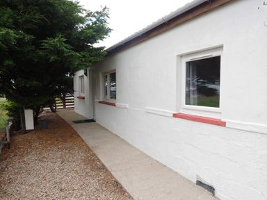 A white exterior wall of a building with windows and a door next to a gravel and concrete path with trees on the side at Braemore Chalet Tarbert Isle Of Harris