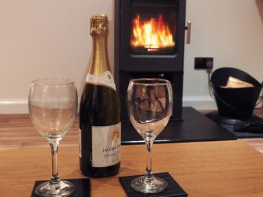 A bottle of sparkling wine and two empty glasses on a wooden table with a lit fireplace in the background at Braemore Chalet in Tarbert Isle Of Harris