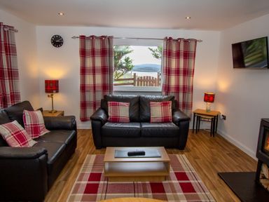 A living room with two black leather sofas red and white plaid cushions and curtains a coffee table a wall mounted television and a fireplace at Braemore Chalet in Tarbert Isle Of Harris