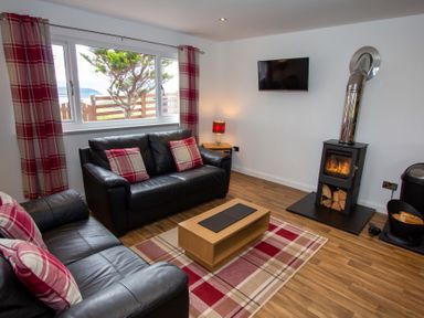 A living room with black leather sofas plaid cushions and curtains a wood burning stove a wall mounted TV and a wooden coffee table at Braemore Chalet Tarbert Isle Of Harris