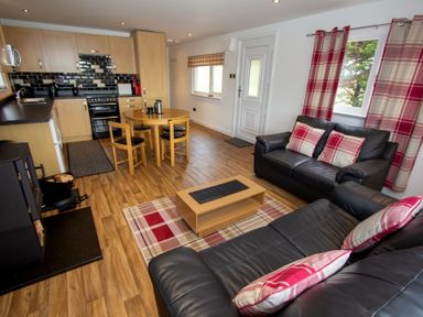 An open plan living room and kitchen with black leather sofas wooden dining table and kitchen cabinets at Braemore Chalet Tarbert Isle Of Harris