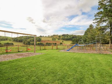 A fenced outdoor playground area with swings and a slide surrounded by green grass and hills at Pen Bont Home Farm in Upper Chapel near Builth Wells