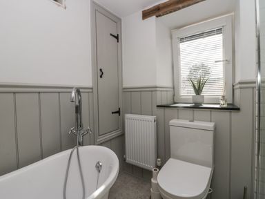 A bathroom with a bathtub faucet a toilet a small cabinet a radiator a window with blinds and a plant on the sill at Style Cottage in Keswick