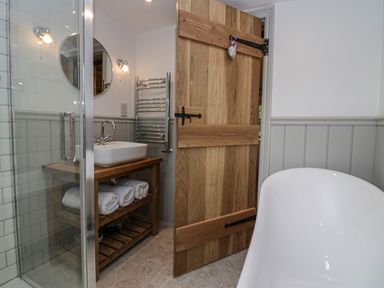 A bathroom with a wooden door a white bathtub a sink on a wooden stand with towels and a glass shower at Style Cottage in Keswick