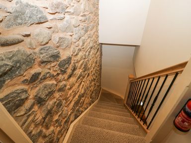 A carpeted staircase with a stone wall on one side and a wooden handrail with metal balusters on the other at Style Cottage in Keswick