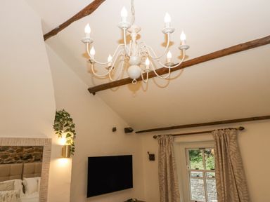 A ceiling with wooden beams and a chandelier above a mounted TV and window with curtains at Style Cottage in Keswick