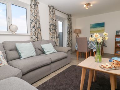 A living room with a gray sofa, light blue cushions, a wooden coffee table with flowers and snacks, dining table with chairs, lamp and bookshelf at Atlantic Reach in Marazion