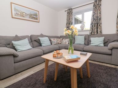 A living room with a grey corner sofa with cushions a wooden coffee table with flowers hot cross buns and a book at Atlantic Reach in Marazion