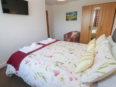 A bedroom with a floral bedspread a wall-mounted television a wooden wardrobe and a checkered armchair at Atlantic Reach in Marazion