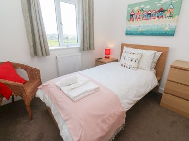 A bedroom with a single bed a pink throw towels on bed a wooden chest of drawers a wicker chair with a red cushion a bedside table with a red lamp and a window with curtains at Atlantic Reach in Marazion