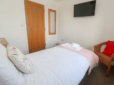 A bedroom with a bed with white and pink bedding a wooden door a wall mirror a flat screen TV and a wicker chair with a red cushion at Atlantic Reach in Marazion