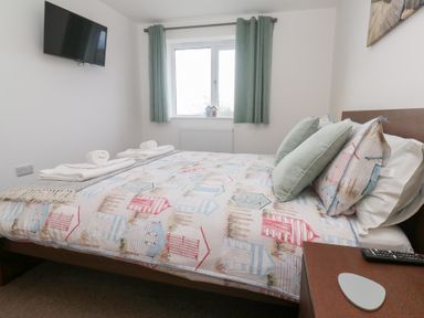 A bedroom with a bed covered in patterned bedding and two pillows a mounted tv a window with curtains and a bedside table at Atlantic Reach in Marazion