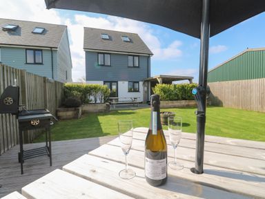 A backyard with a table holding a bottle and two glasses under an umbrella with two houses in the background at Atlantic Reach in Marazion