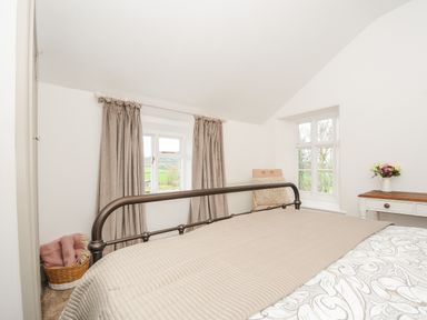 A bedroom with a metal bed frame a basket with blankets beige curtains a chair and a table with flowers at Skylarks No Mans Land near Looe